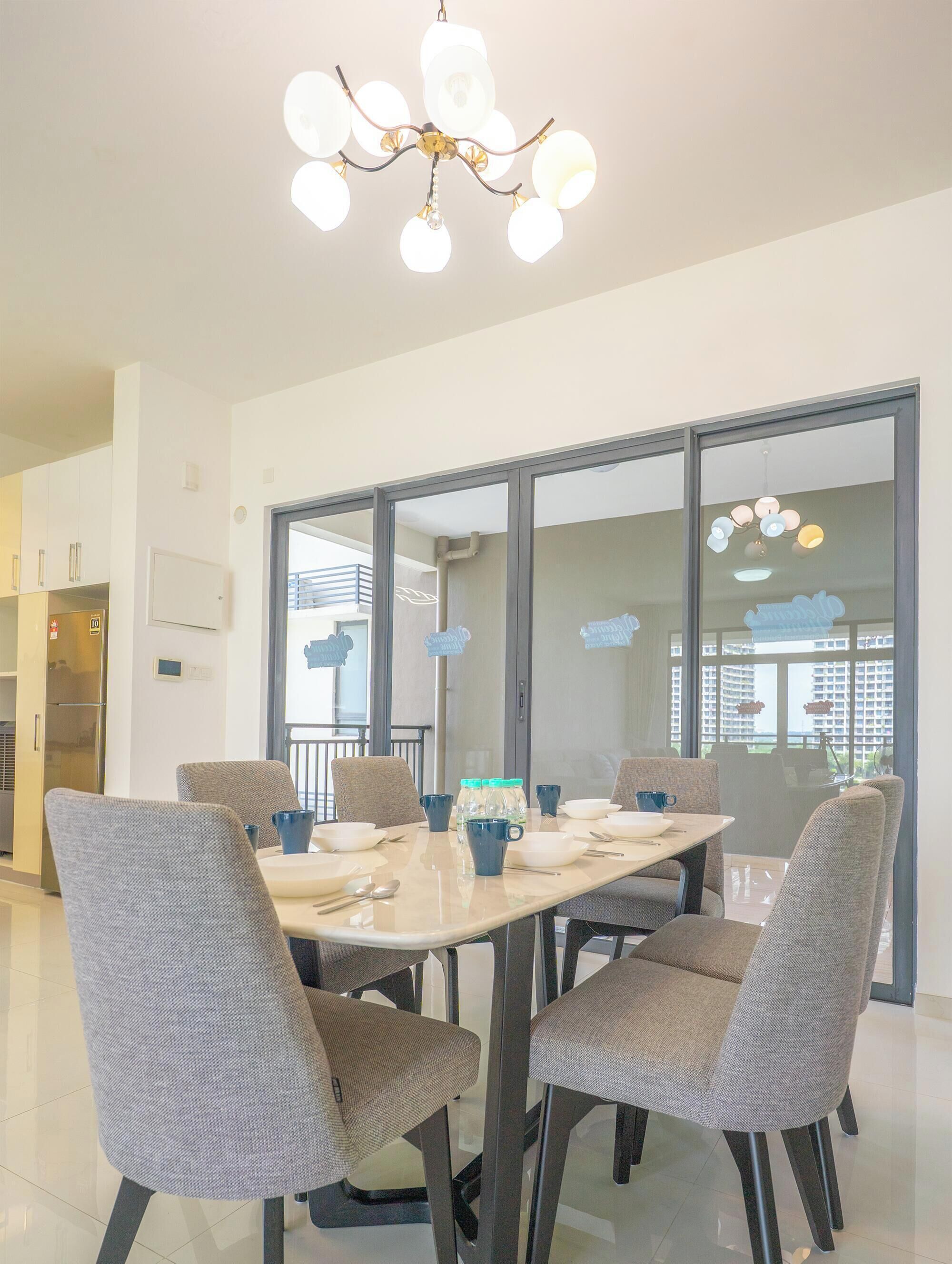 A dining area with a table set for six, surrounded by six grey chairs. A chandelier hangs from the ceiling, and the room has large windows with a view of buildings outside.