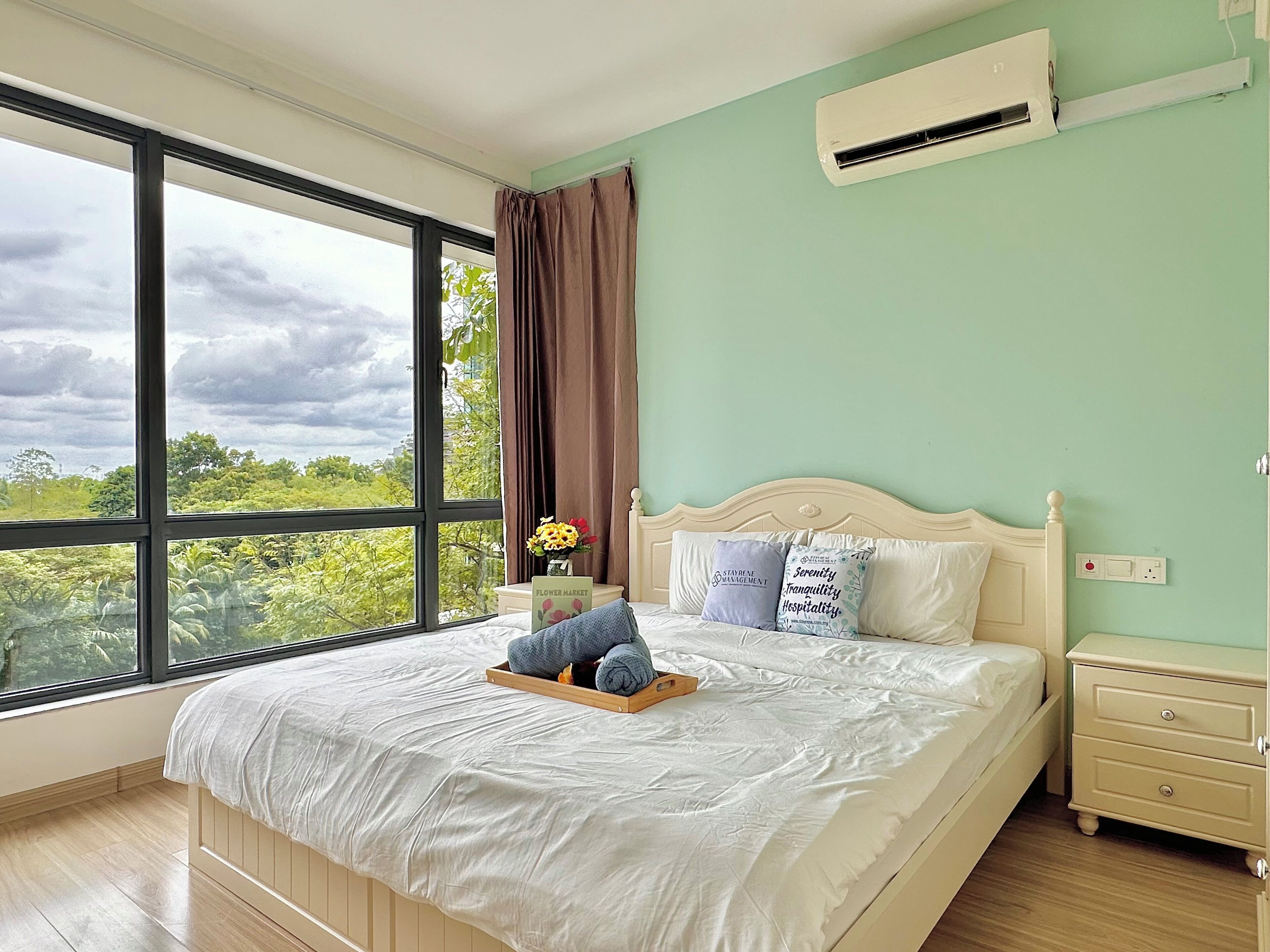 A bedroom with a large window, a bed with a wooden headboard, a nightstand, and a small table with a vase of flowers.