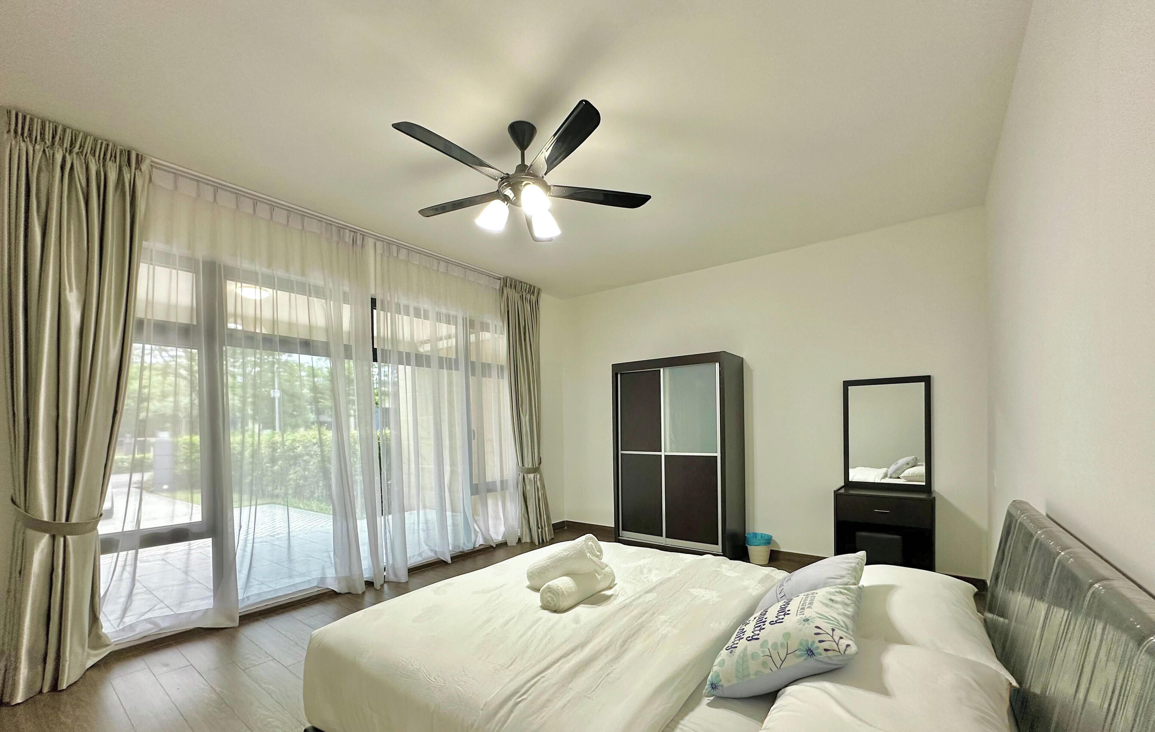 A hotel room with a bed, a ceiling fan, a large window with curtains, a wardrobe, and a bedside table.