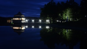 A lakeside building with a gazebo-like structure, illuminated by multiple lights, reflecting on the water.