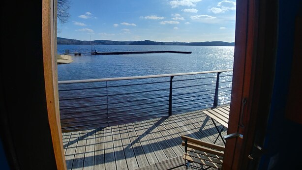 A wooden deck with a bench, overlooking a calm lake with a dock and distant hills.
