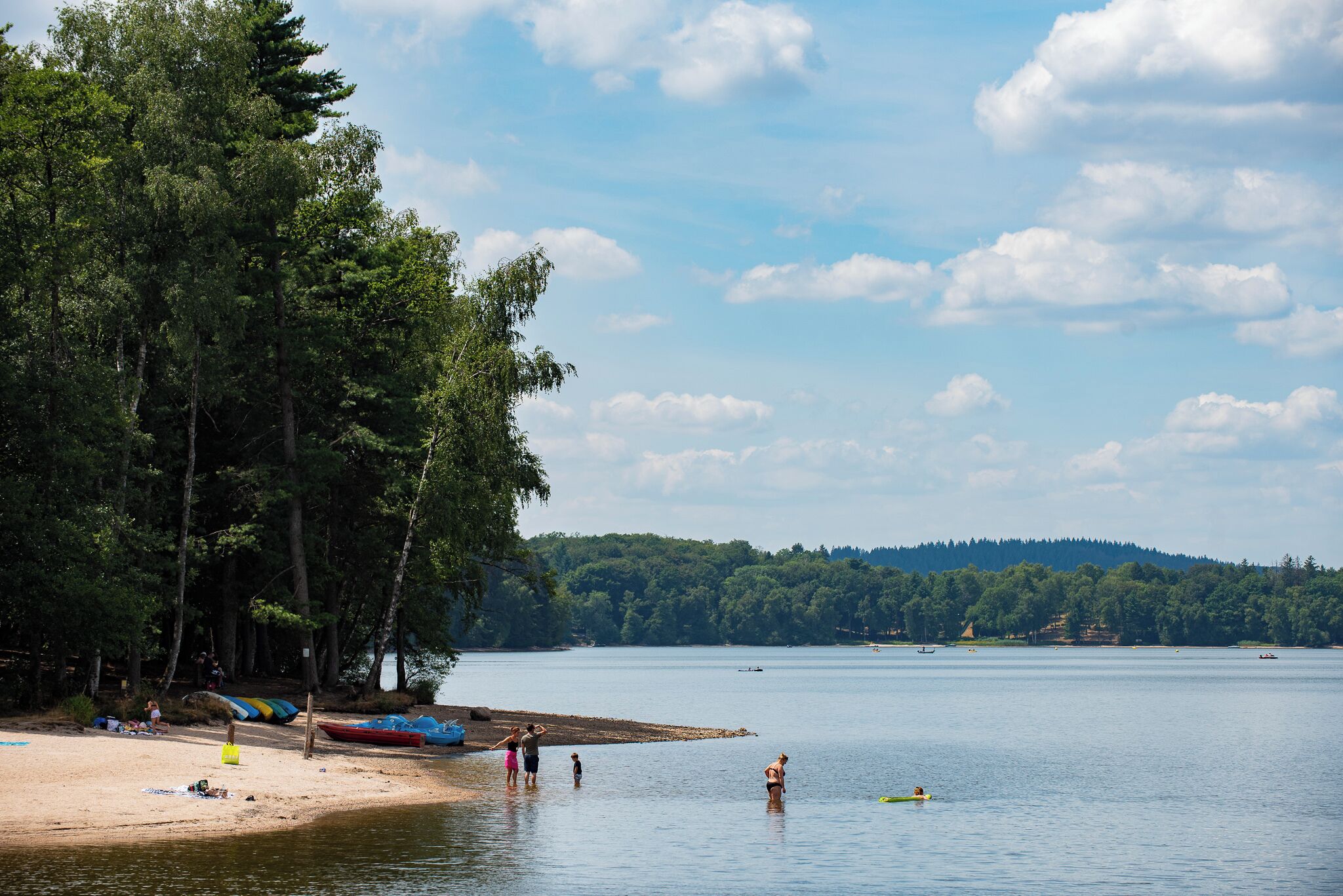 A lakeside beach with people swimming and standing on the shore, surrounded by trees and boats.