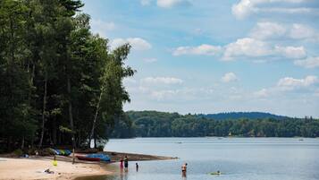 A lakeside beach with people swimming and standing on the shore, surrounded by trees and boats.