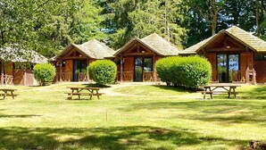 A row of wooden cabins with picnic tables in a grassy area surrounded by trees.
