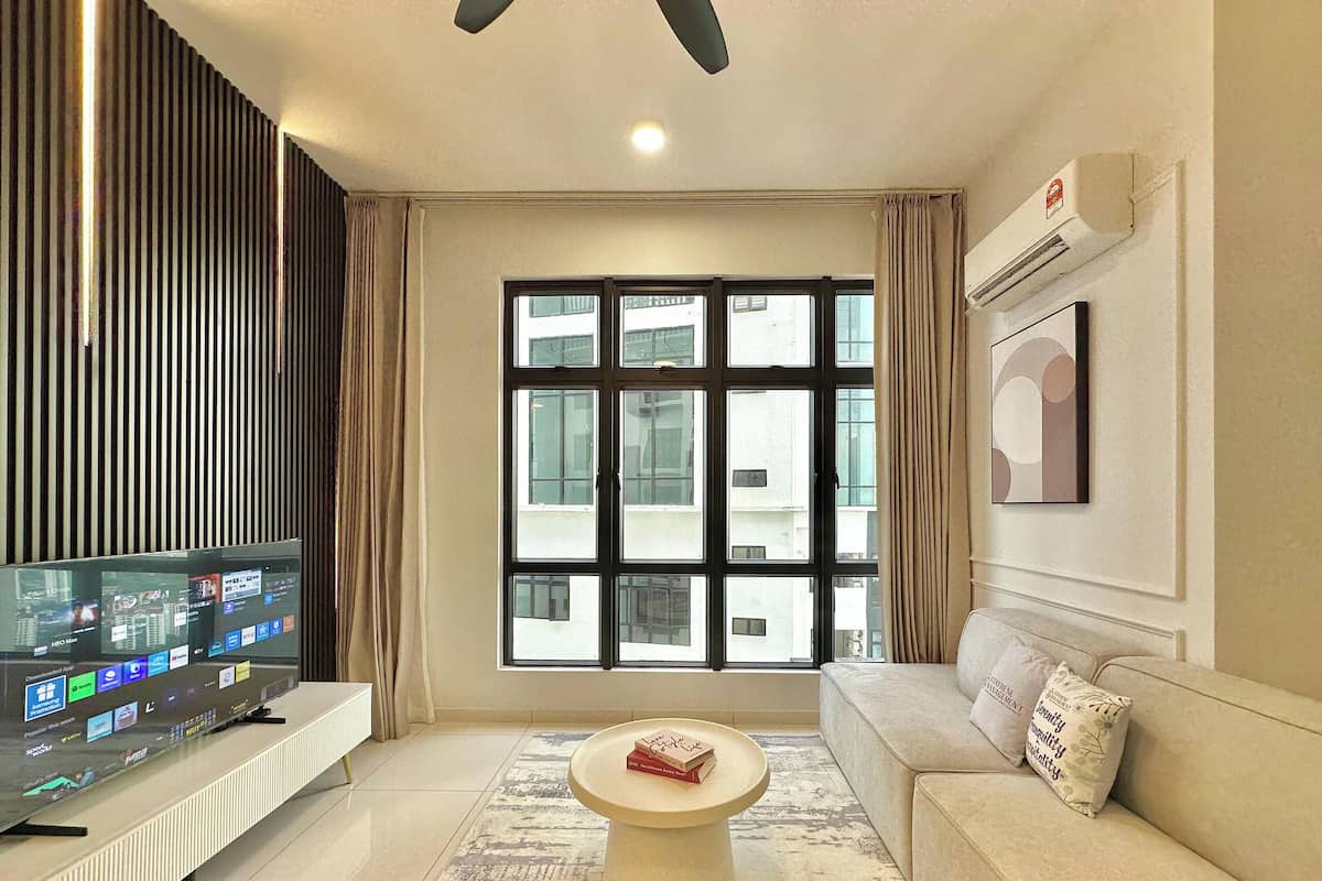 A modern living room with a flat-screen TV, a round coffee table, a sofa, a large window with blinds, and an air conditioning unit.