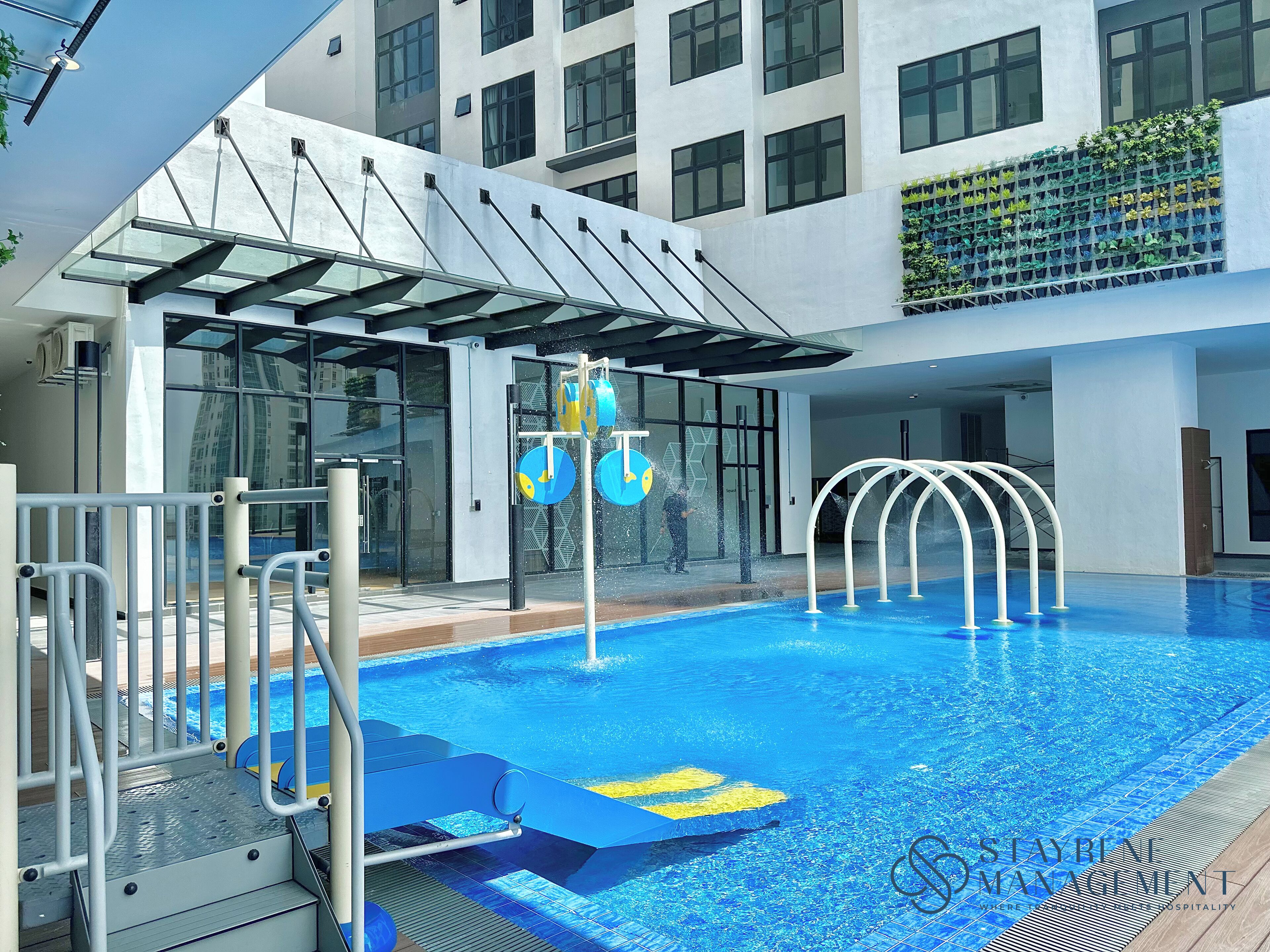 Ein moderner Hotelpoolbereich mit einer gläsernen Struktur, einer Wasserspielen und Blick auf die Außenfassade des Gebäudes.