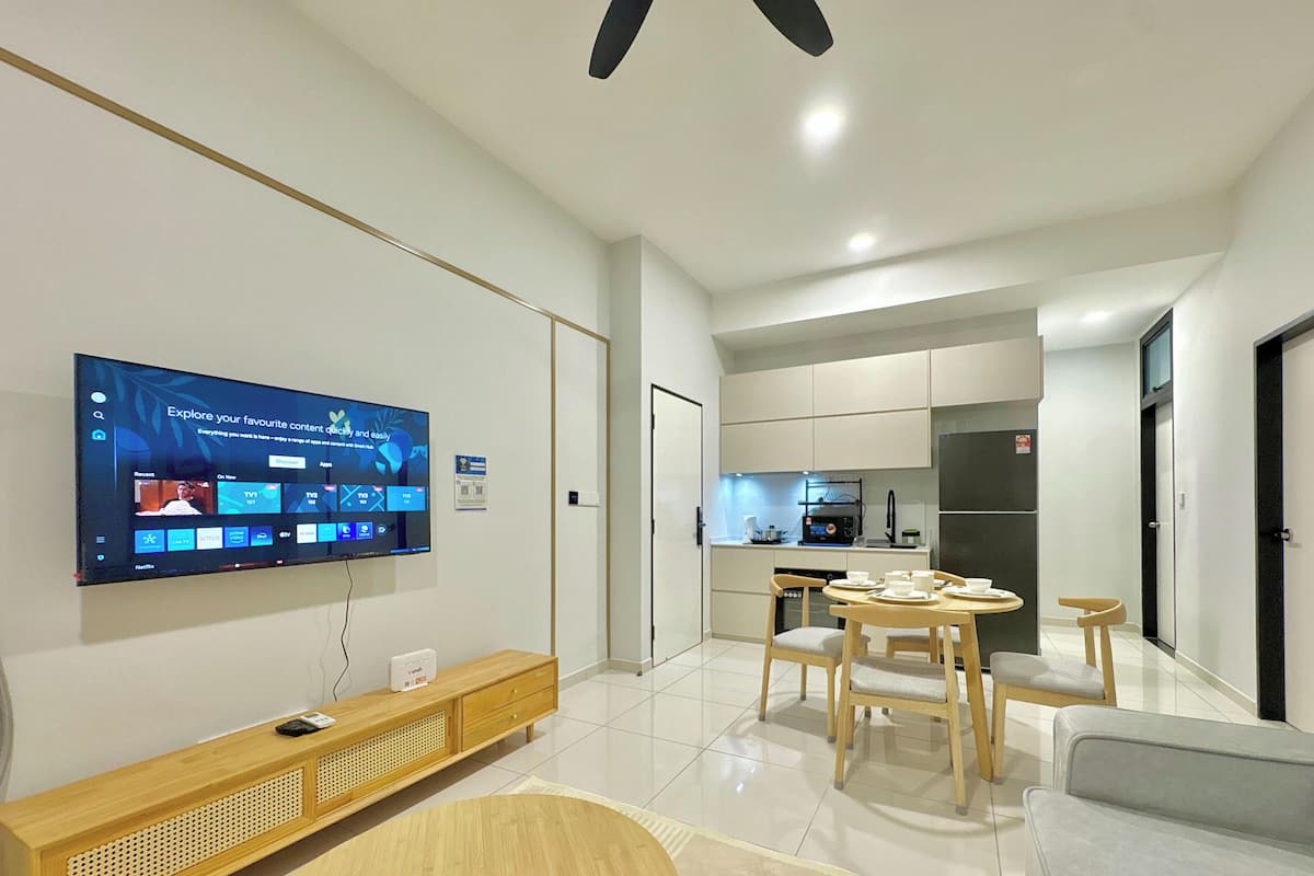 A modern living room with a flat-screen TV, a wooden entertainment unit, a dining area with a table and chairs, and a kitchen in the background.