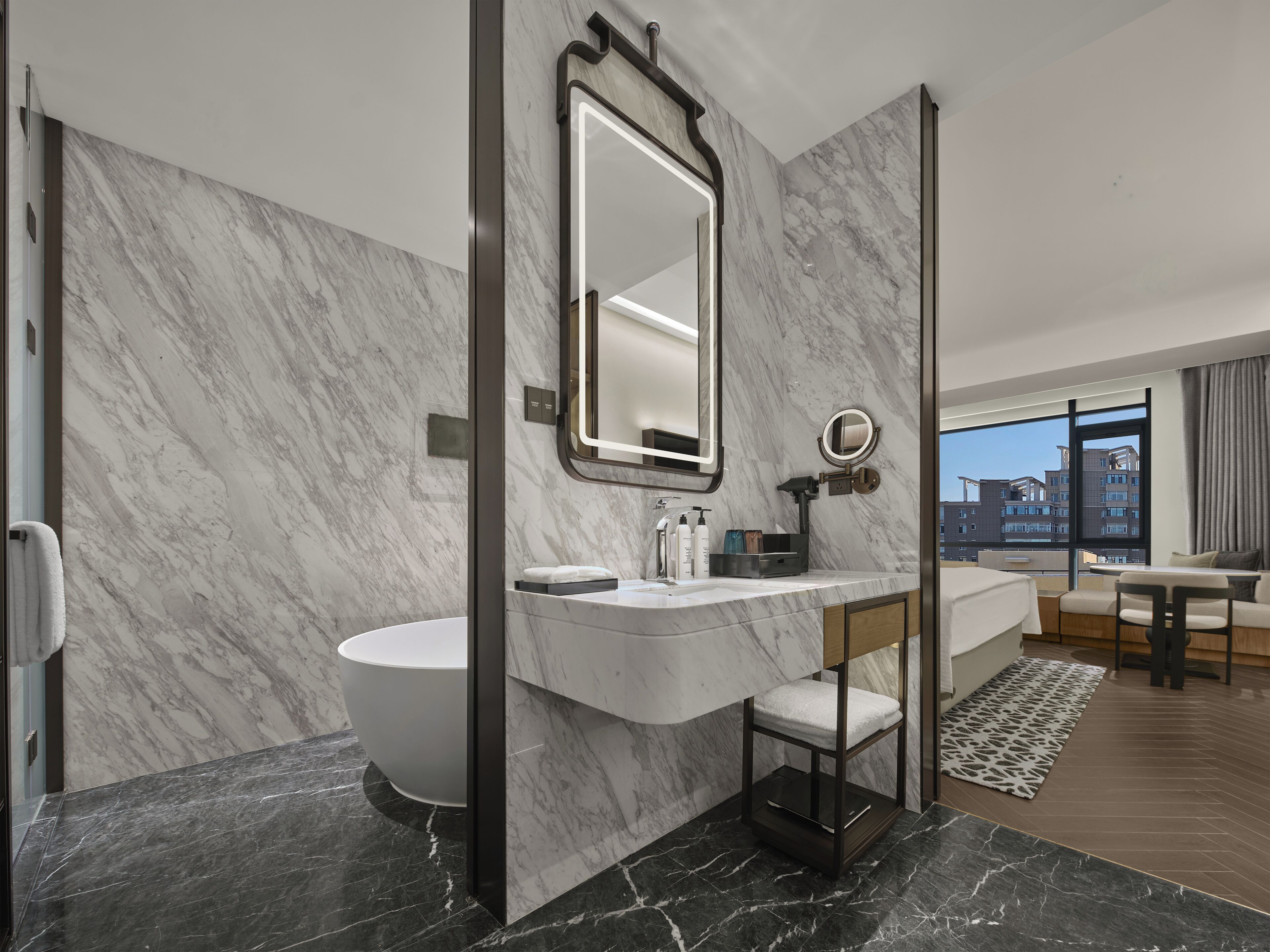 A modern bathroom with a marble wall, a large mirror, a bathtub, and a vanity with toiletries.