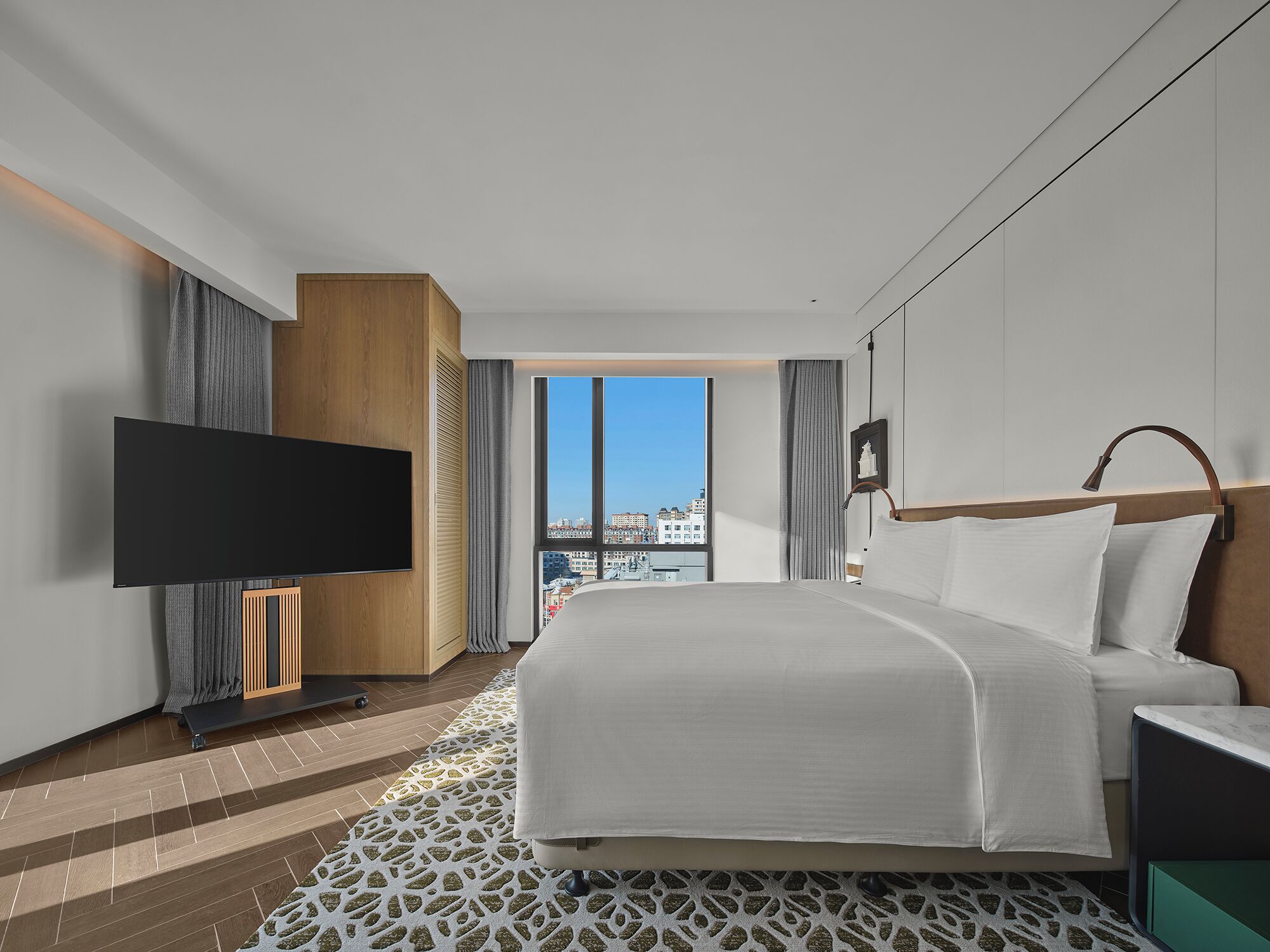 Une chambre d’hôtel moderne dotée d’un grand lit, d’un téléviseur à écran plat, d’une vue sur la ville et d’un sol à motifs.