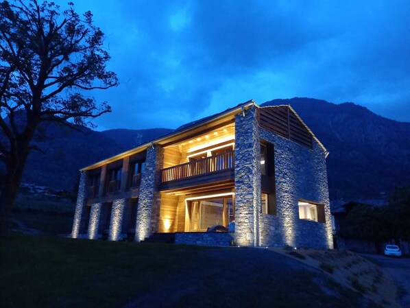 A two-story stone house with a balcony, set against a mountainous backdrop.