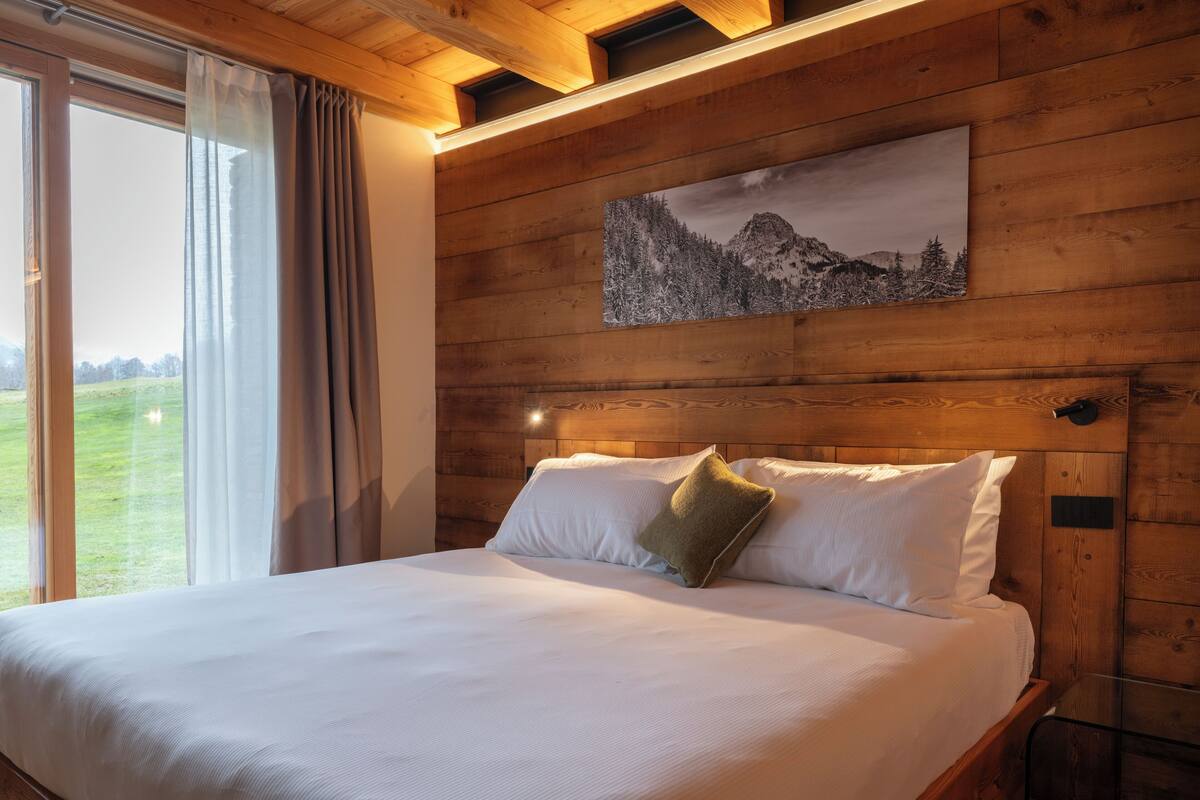 Ein Schlafzimmer mit Holzwandverkleidung, einem großen Bett, einem Gemälde von einer Berglandschaft und Blick auf ein grünes Feld durch ein Fenster.