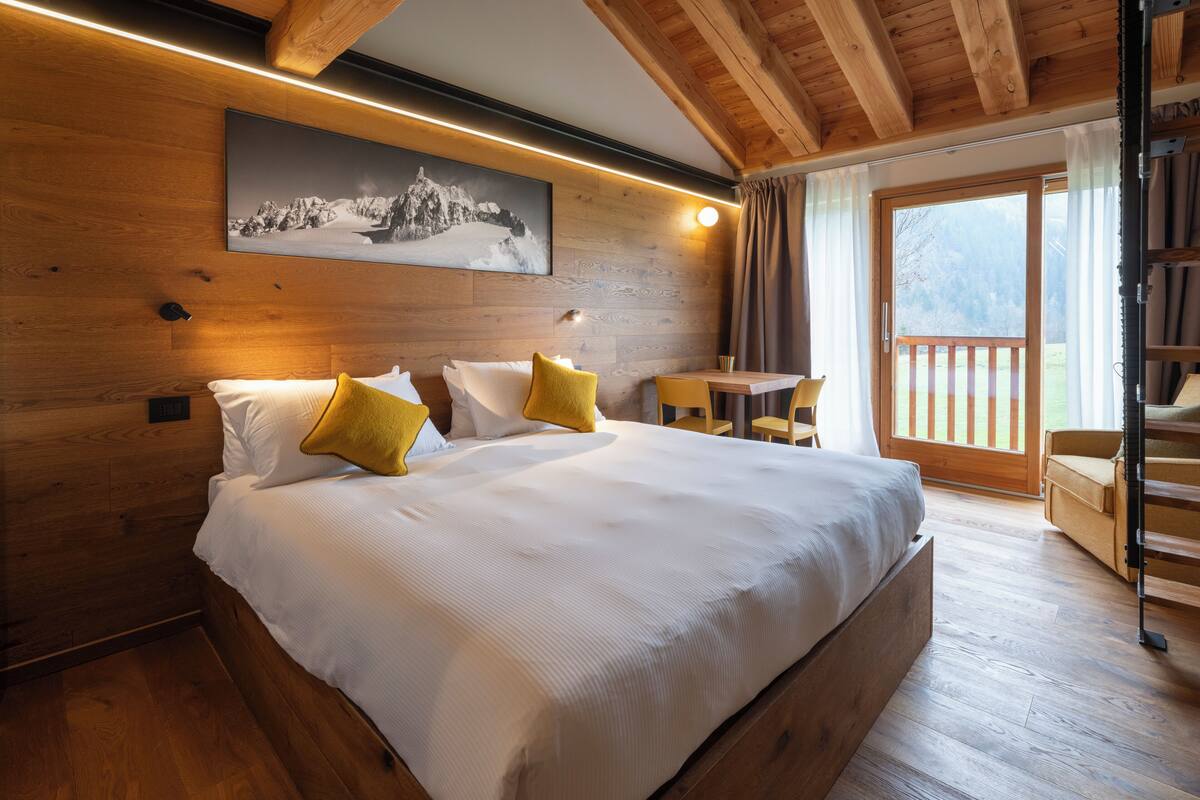 Ein Schlafzimmer mit Holzbalkendecke, einem Bett mit weißer Bettwäsche und senffarbenen Kissen, einem Tisch mit Stühlen und einem Fenster mit Blick ins Freie.