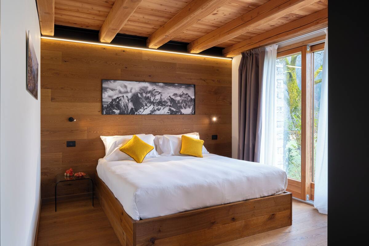 Ein Bett mit Holzrahmen, weißer Bettwäsche und zwei gelben Kissen, ein Holz-Kopfschild, ein an der Wand hängendes Bild und ein Fenster mit Vorhängen.