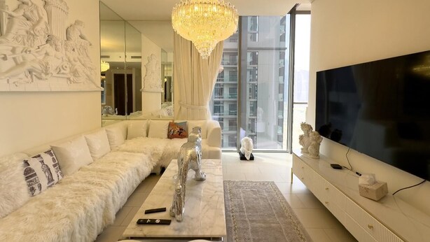 A modern living room with a large flat-screen TV, a white sofa, a marble coffee table, and a chandelier. - Manzil - 1BR in Sobha with Skyline Views (Dubai)