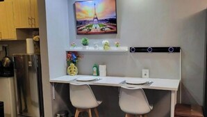A compact kitchen with a white countertop, two chairs, a stainless steel refrigerator, and a wall-mounted TV above a shelf with decorative items. - Cozy 1-bedroom condo with AC in charming Muntinlupa (Muntinlupa)