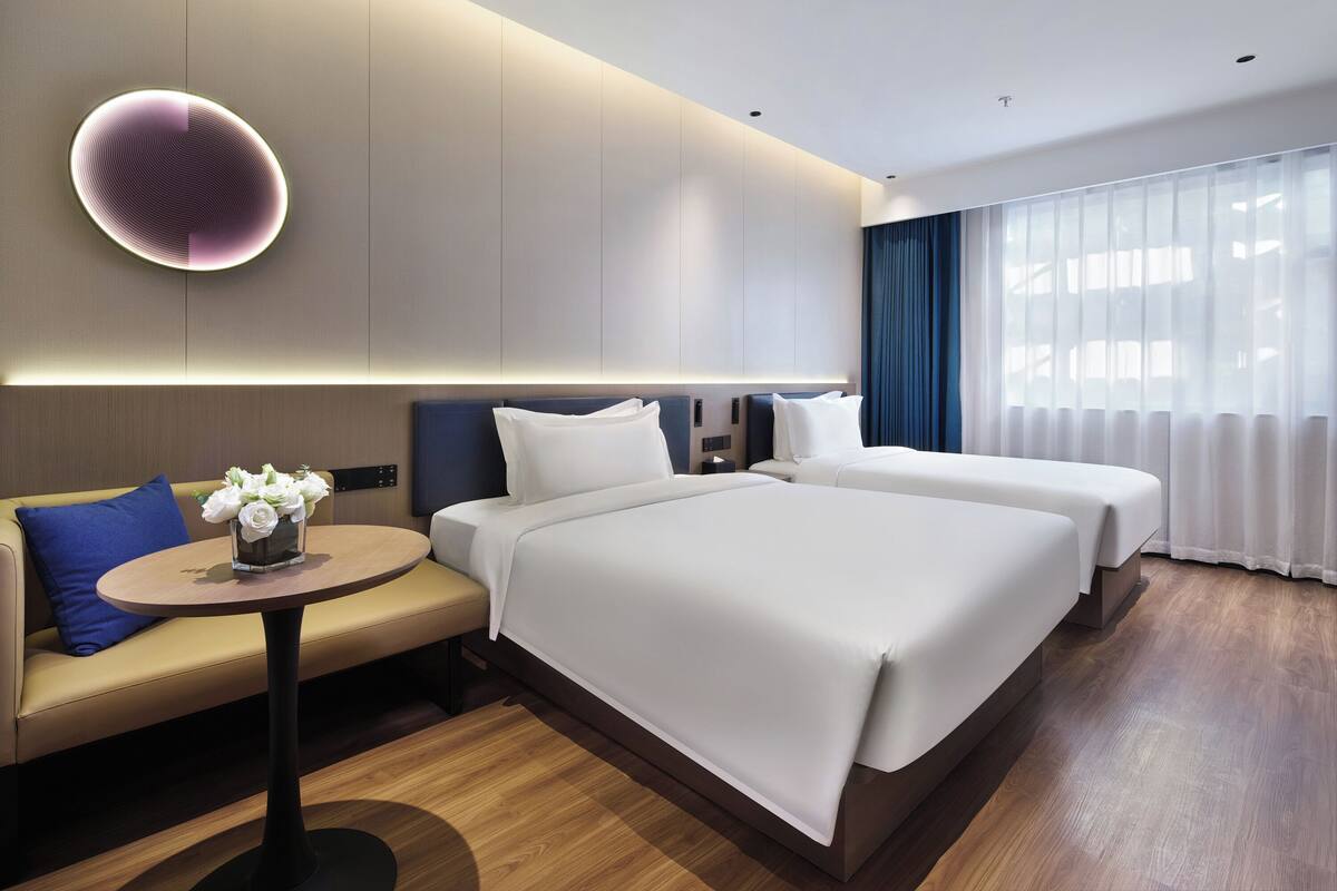 A modern hotel room with a large bed, a sofa, a small round table, and a wall-mounted light fixture.