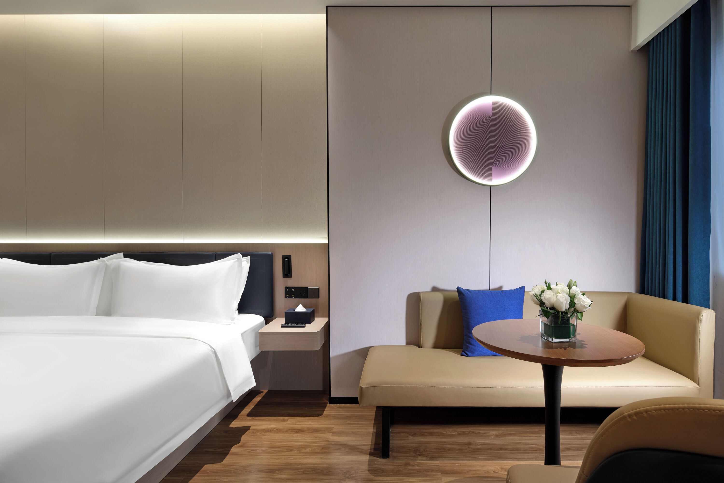 A modern hotel room with a bed, a sofa, a small table with a vase of flowers, and a wall-mounted light fixture.
