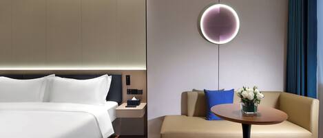 A modern hotel room with a bed, a sofa, a small table with a vase of flowers, and a wall-mounted light fixture.