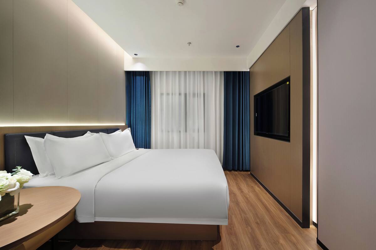 A modern hotel room with a large bed, a bedside table with a vase of flowers, a flat-screen TV mounted on the wall, and a large window with curtains.
