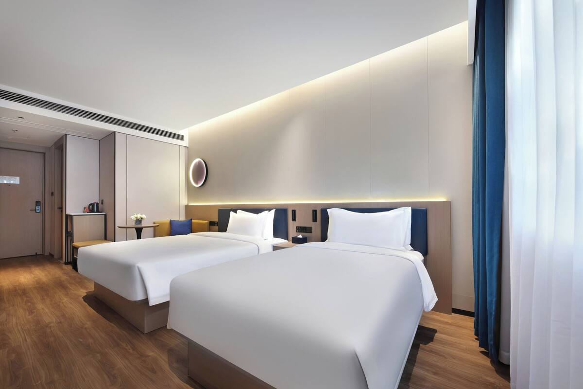 A modern hotel room with two beds, a small table with a vase, and a wall-mounted control panel.