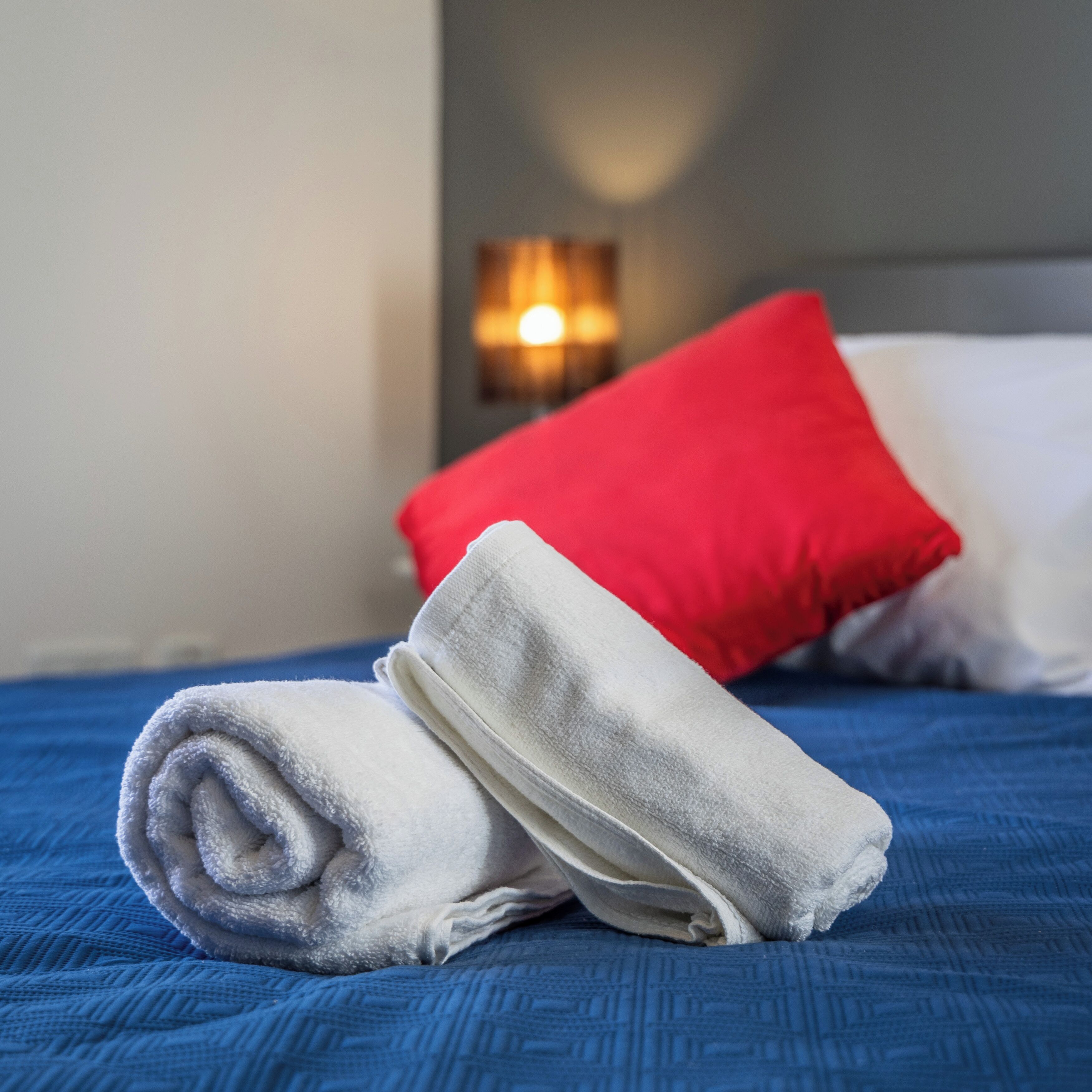 Un lit bien fait, avec une couverture bleue, une serviette blanche roulée et un oreiller rouge.