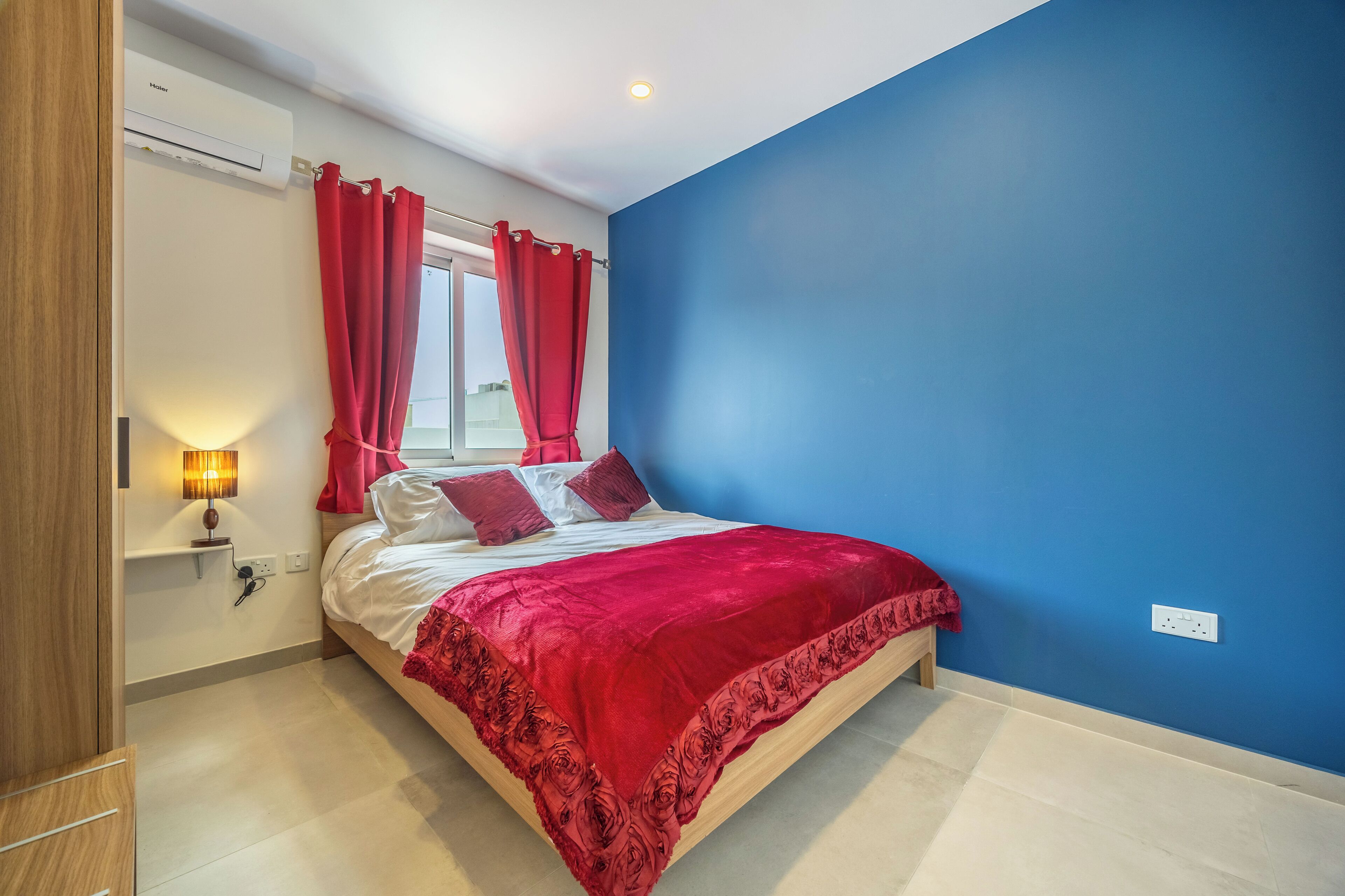 Un dormitorio con cama, cortinas rojas, una mesita de noche con lámpara y pared azul.