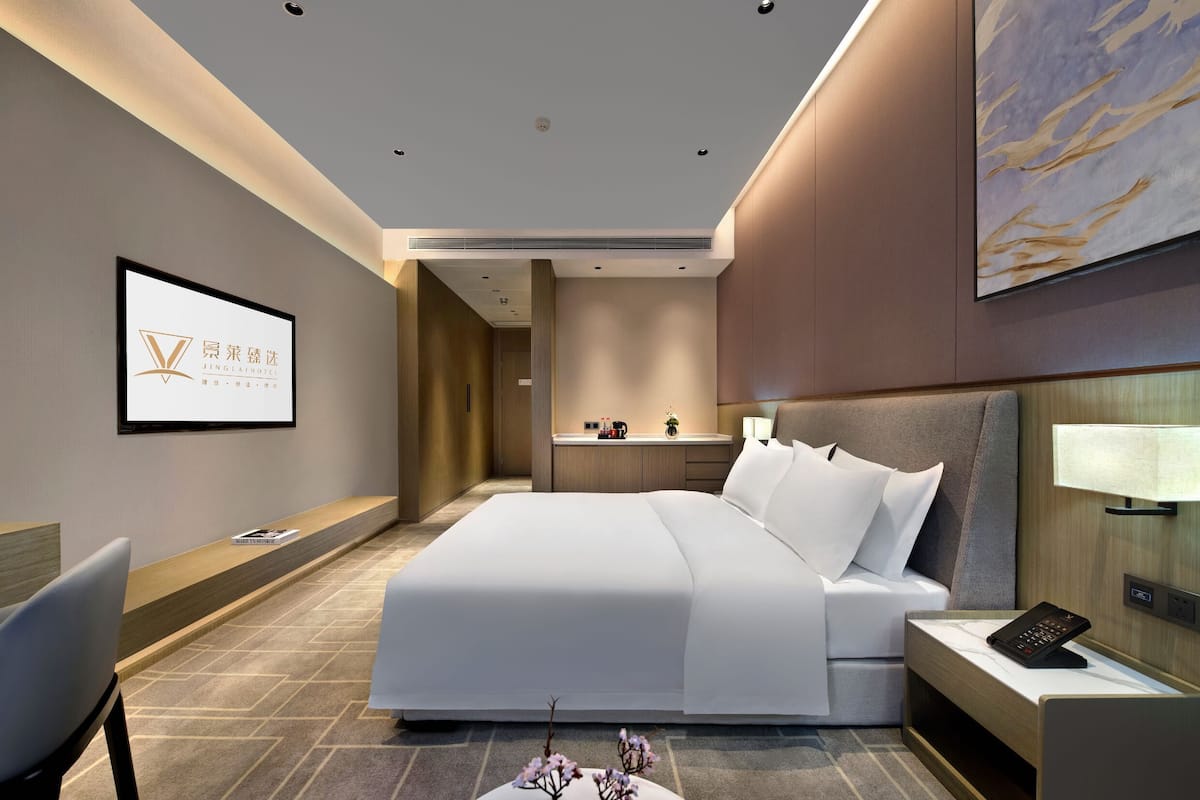 Una habitación de hotel moderna con una cama grande, un escritorio, una silla, una mesita de noche con lámpara, un televisor y un cuadro en la pared.