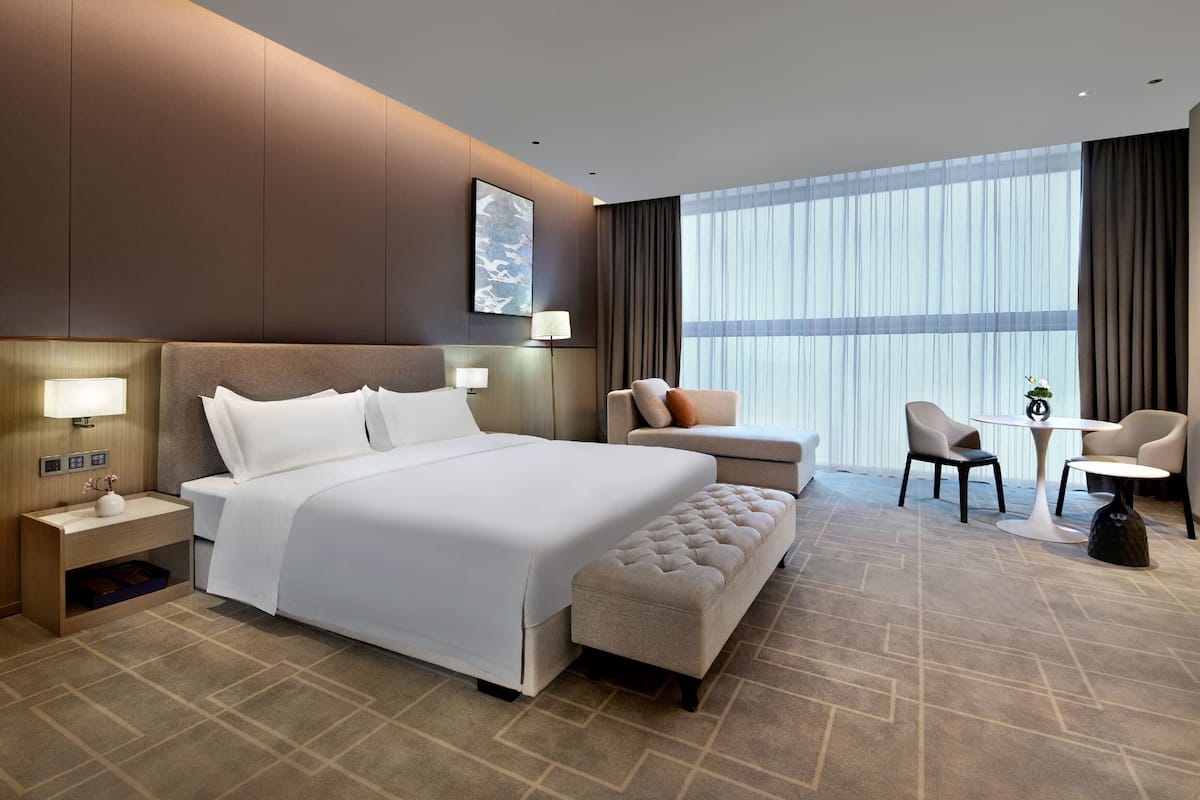Habitación de hotel moderna con una cama grande, una mesita, dos sillas, un banco y un ventanal con cortinas.