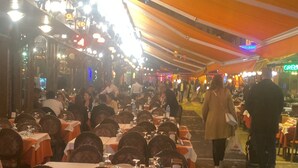 Uma área externa para refeições com mesas e cadeiras sob um toldo grande, cercada por outros restaurantes.
