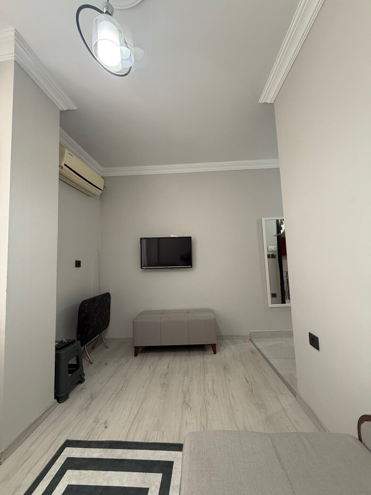 A minimalist living room with a flat-screen TV mounted on the wall, a grey sofa, a small black stool, and a black and white rug.