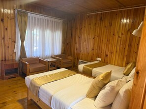 A room with wooden walls, two beds with white and gold bedding, a wooden desk, a chair, and a window with curtains.