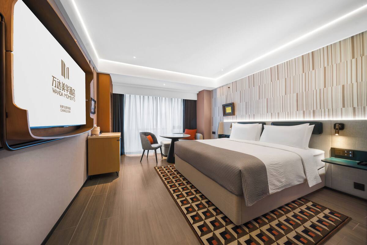 A modern hotel room with a large bed, a desk, a chair, and a TV mounted on the wall.