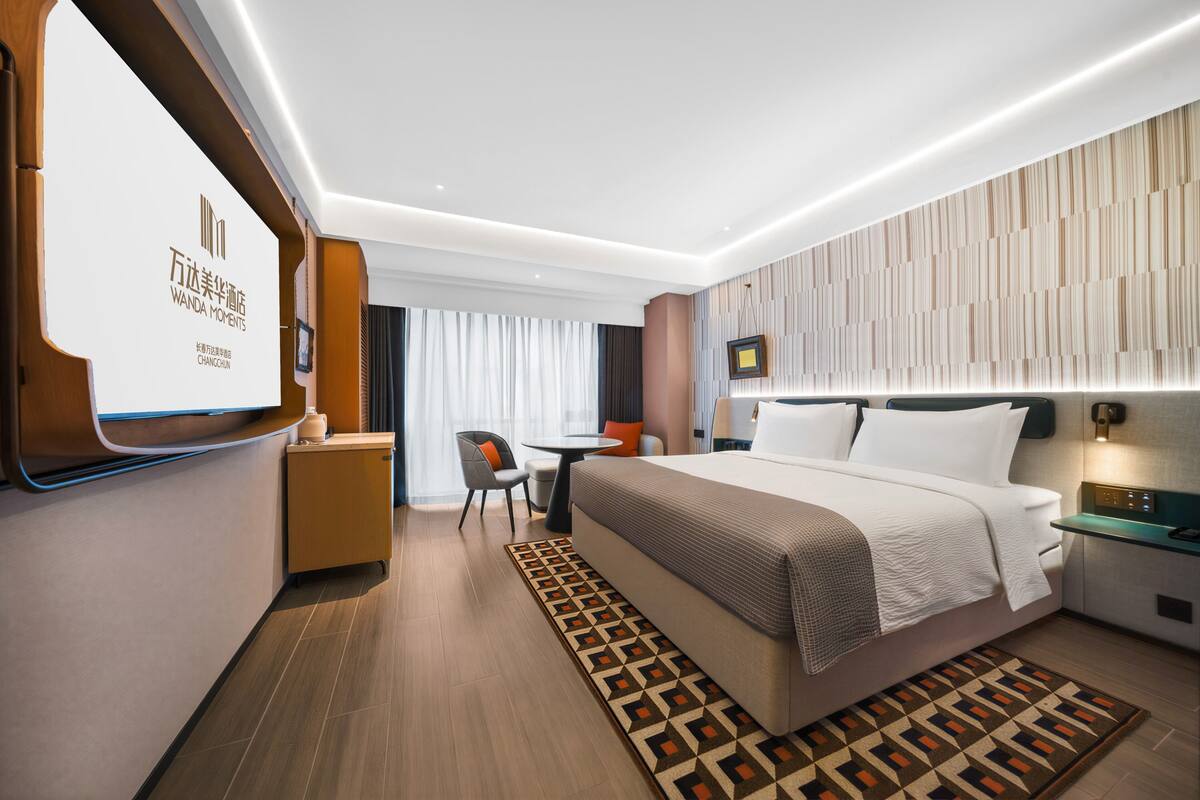 A modern hotel room with a large bed, bedside table, and a desk with a chair.