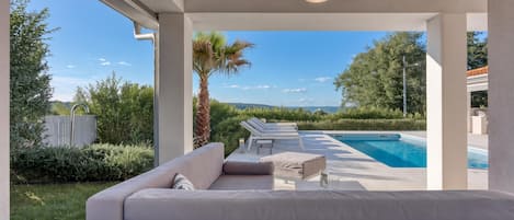 A poolside area with a sofa and lounge chairs, a palm tree, and a view of the pool and landscape.