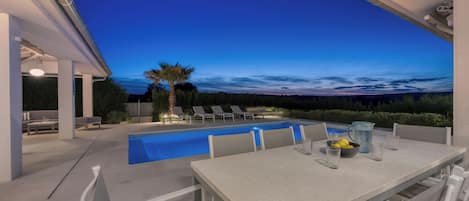 An outdoor pool area with a table and chairs, a palm tree, and a view of the sky.
