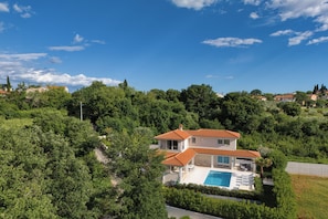 Una casa de dos pisos con piscina, rodeada de abundante vegetación y árboles.