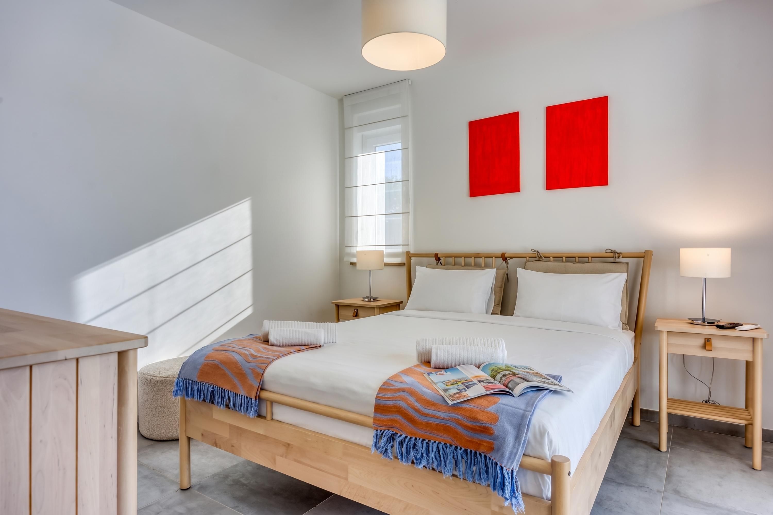 Een moderne slaapkamer met een houten bed, nachtkastjes, een raam en twee rode kunstwerken aan de muur.
