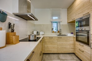 A modern kitchen with wooden cabinetry, a stainless steel range hood, a built-in oven, and a black induction cooktop. - Pool Villa Ora in Nature (Marcana)