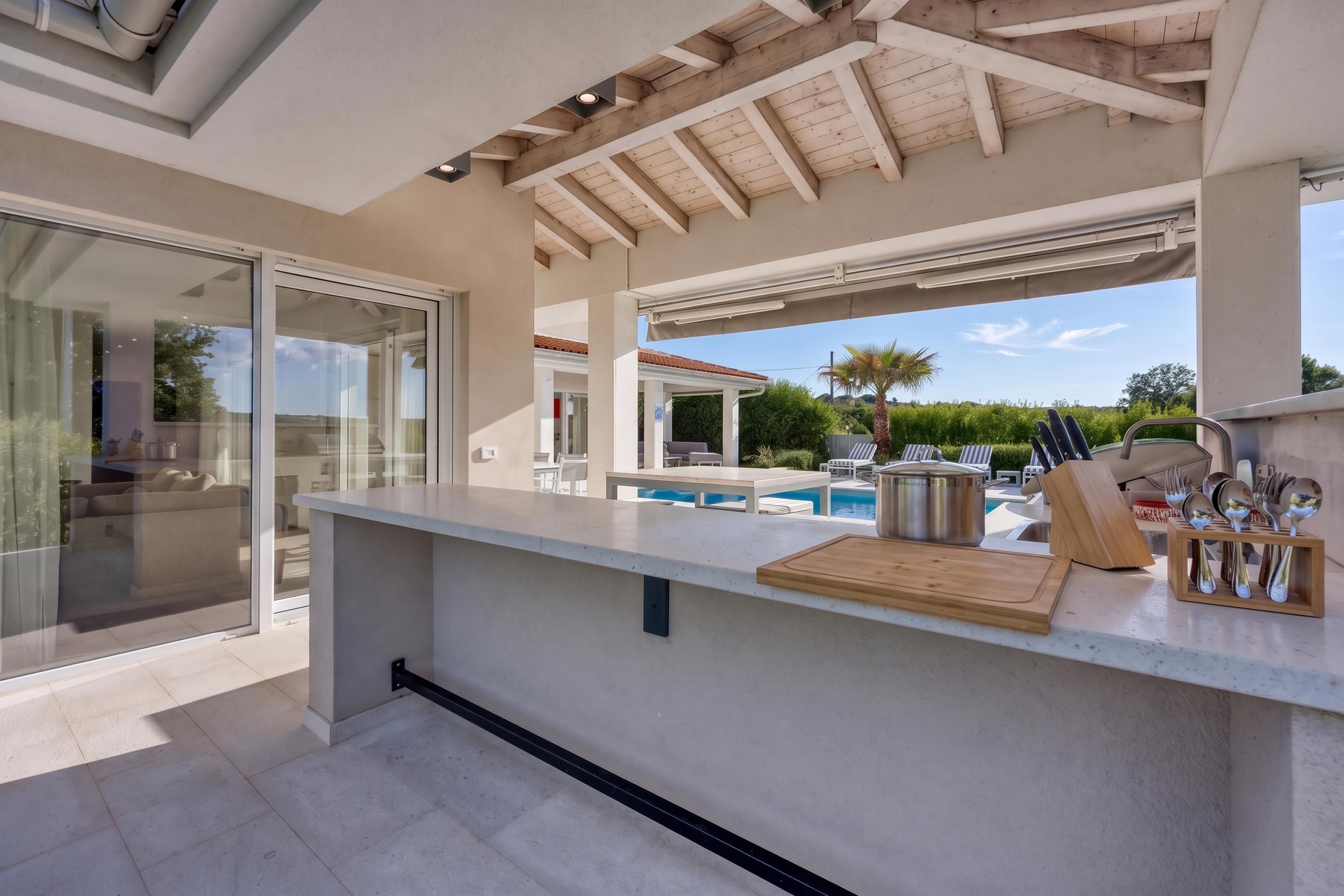 A modern outdoor kitchen with a pool, a dining area, and a glass door leading to a poolside lounge.