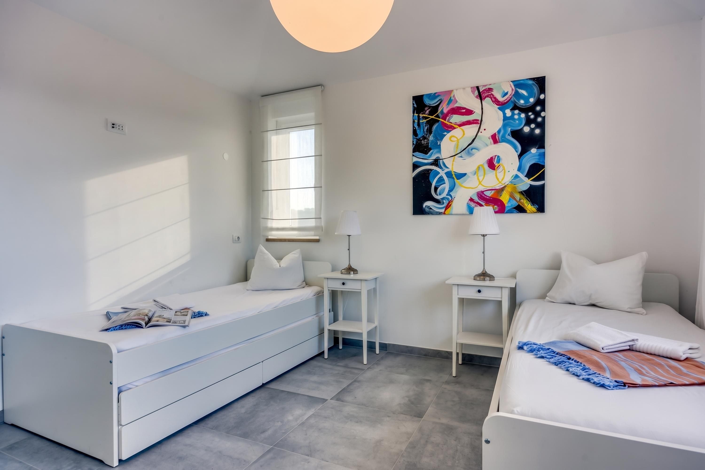 A modern bedroom with two beds, a window with blinds, a painting on the wall, and a small table with a lamp.
