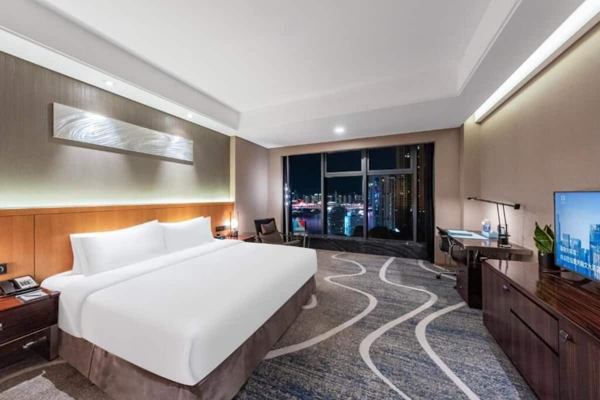 Une chambre d’hôtel avec un grand lit, un bureau avec un ordinateur, une télévision et une vue sur la ville la nuit.