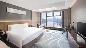 Ein Hotelzimmer mit einem großen Bett, einem Schreibtisch, einem Fernseher und Blick auf eine Stadtkulisse durch ein großes Fenster.