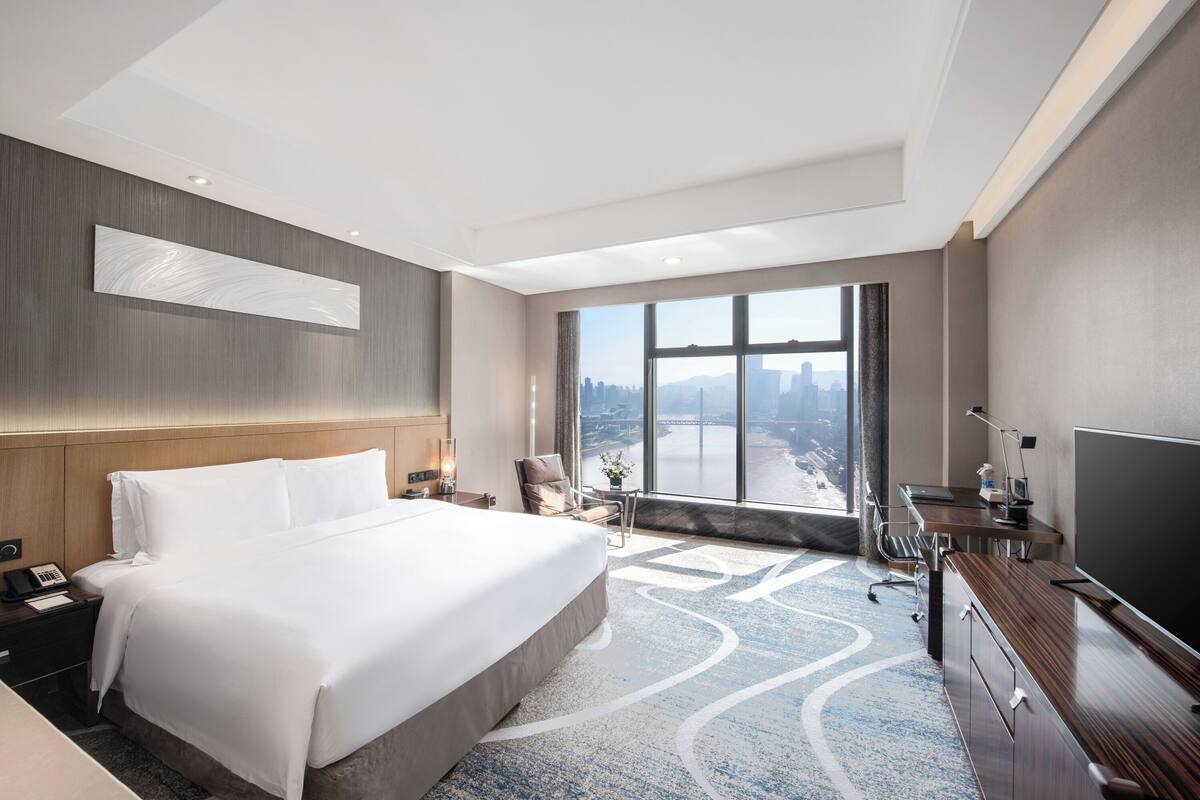 Une chambre d’hôtel avec un grand lit, un bureau, une télévision et une vue sur le paysage urbain par une grande fenêtre.