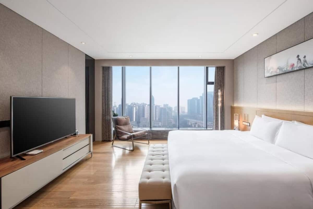 Une chambre d’hôtel moderne dotée d’un grand lit, d’un téléviseur à écran plat, d’un coin salon avec un fauteuil et offrant une vue sur la ville grâce à de grandes fenêtres.