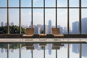 Ein moderner Poolbereich im Innenbereich mit zwei Holzliegestühlen und einem kleinen Tisch, große Fenster mit Blick auf die Stadt.