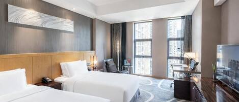 A hotel room with two beds, a desk, a TV, and a large window with a city view.
