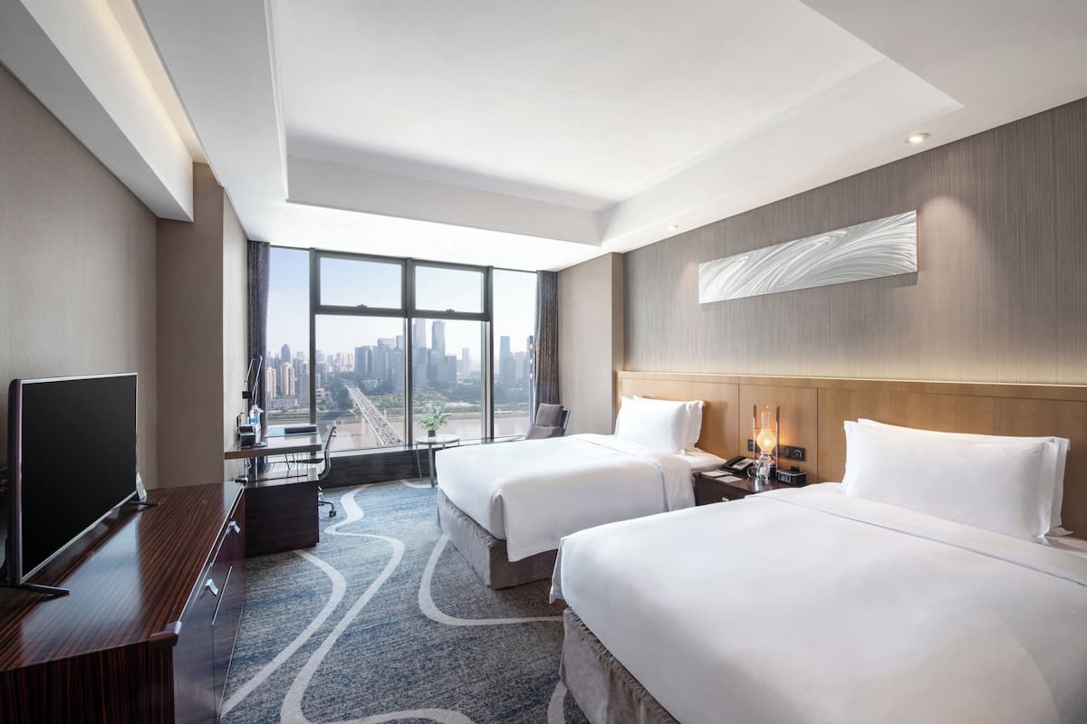 Une chambre d’hôtel avec deux lits, un bureau avec une télévision, une grande fenêtre donnant sur la ville et un décor moderne.
