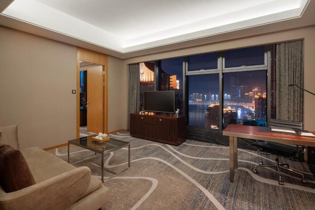 Una habitación de hotel moderna con un amplio ventanal que ofrece vista a la ciudad de noche, un televisor de pantalla plana, un sofá, una mesa de centro y un escritorio con una silla.