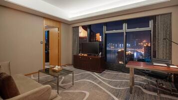 Una habitación de hotel moderna con un amplio ventanal que ofrece vista a la ciudad de noche, un televisor de pantalla plana, un sofá, una mesa de centro y un escritorio con una silla.