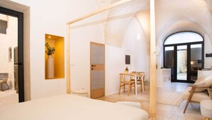Ein Schlafzimmer mit einem großen Bett, einem Holztisch und einem Stuhl. Ein kleiner Tisch beherbergt eine Vase und eine Pflanze. Der Raum ist weiß gestrichen und hat einen Fliesenboden.